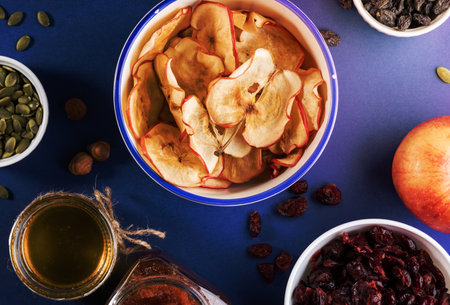Close-up ceramic cup with homemade apple chips surrounded by healthy snacks and jars of jam and honey on blue background. Natural fruit healthy snack. Top view, flat lay.の写真素材