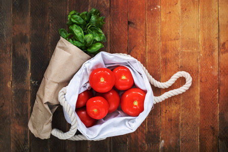 Red tomatoes in white linen bag and green Basil in paper packaging on wooden table. Plastic free concept, ecologic pack. Copy space, top view.の写真素材