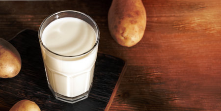 Potato milk glass close up on wooden serving board on wooden background. Lactose-free alternative milk. Organic healthy drink. Vegetable based milk replacer. Banner with copy space. Selective focus.の写真素材