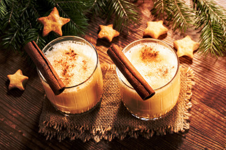 Two glasses of homemade eggnog with cinnamon, fir branches, cookies stars on burlap on rustic wooden table. Traditional Christmas drink. Holiday cozy image. Low key. Back Lit with shadows. Top view.の写真素材