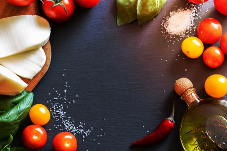 Ingredients for traditional Italian Caprese salad: red and yellow tomatoes, mozzarella cheese, salt, olive oil and green Basil are lying in circle on dark shale board with copy space in the middle. Top view, flat lay.の写真素材