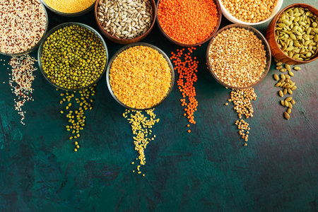Top view to round glass and wooden bowls with healthy cereals, beans and seeds and small piles of scattered cereals on textured emerald background. Flat lay. Copy space. Healthy food. Superfoods in cups: red lentils, mung bean, green buckwheat, brown rice, bulgur, quinoa, soya, couscous and seeds of sunflowers and pumpkin.の写真素材