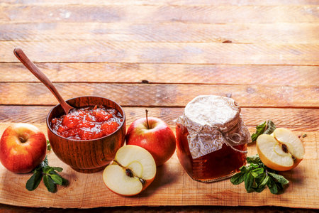 Wooden bowl with spoon and small glass jar closed with homemade paper lid with homemade apple jam and apples and green mint on wooden plank on old rustic wooden table. Natural homemade food. Copy space.の写真素材