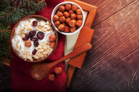 Wooden and ceramic bowls of yogurt with dried cranberries and hazelnuts, spoon and fir twig on red kitchen napkin on tray on wooden table. Top view, copy space.の写真素材