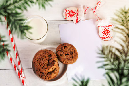 Christmas layout of milk glass, straws, cookies, empty blank sheet, decorative patterned mitts on white wooden table. Milk for Santa. Top view through green fir branches. Horizontal shot. Copy space.の写真素材