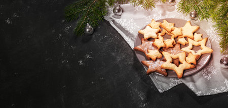 Christmas advertising banner with festive baking. Plate with Xmas cookies of stars shape on grey festive kitchen towel, fir branches with small Xmas balls on black textured backdrop. Copy space.の写真素材