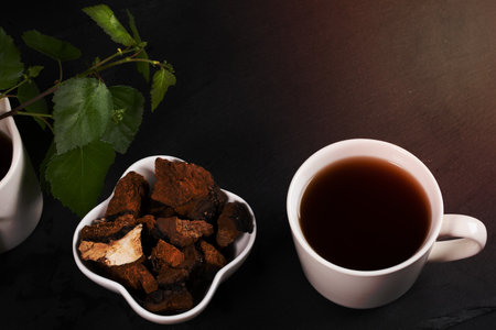 White ceramic cup of healing infusion of birch mushroom chaga, bowl with chaga pieces and birch leaves on black. Chaga mushroom coffee or tea. Trendy healthy superfood for immunity. Copy space.の写真素材