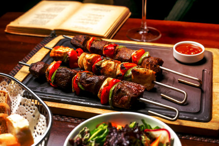 top view of shish kebab with colorful bell peppers served with ketchup and fresh vegetable salad on a black boardの写真素材