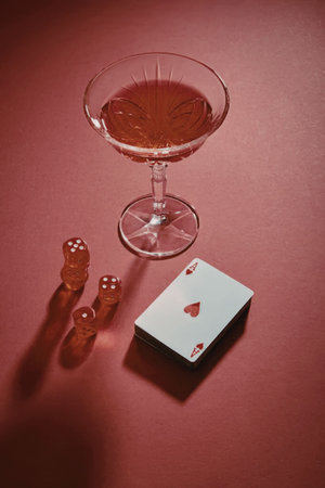 A still life composition featuring a red-colored drink in a glass, a deck of cards, and red game...