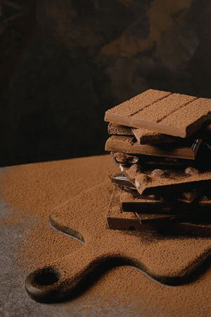 A stack of chocolate bars on a surface covered with cocoa powder, with a dark background.