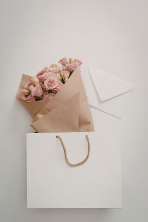A white shopping bag with a brown handle containing pink roses wrapped in brown paper and a white...