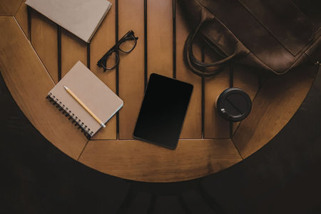 An overhead view of a wooden table with a notebook, glasses, a black tablet, a cup of coffee, and a...