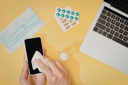 A person cleaning their phone with a white cloth on a yellow surface with a face mask, pills, and a...