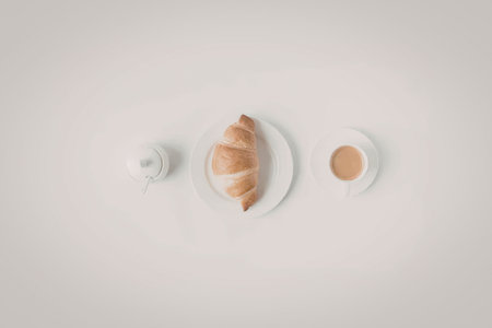 A flaky croissant on a white plate accompanied by a cup of coffee and a small pitcher.