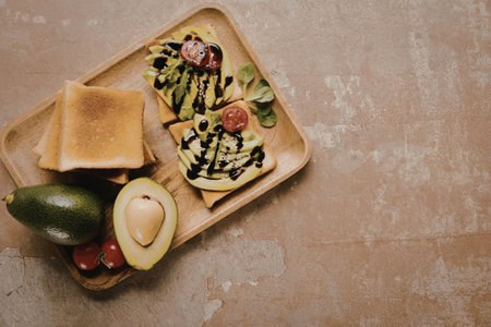 A wooden plate with avocado toast, fresh fruits, and bread on a brown surface