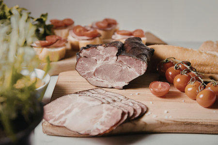 A wooden cutting board with sliced meat, cherry tomatoes, bread, and herbs, arranged for a meal or...