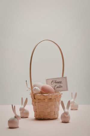 A wicker Easter basket filled with eggs and surrounded by small bunny figurines on a pink surface.