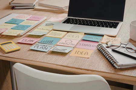 A cluttered desk with sticky notes, laptop, notebook, glasses, and a mug, indicating a workspace for...