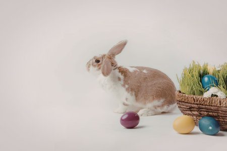 A brown and white Easter bunny sitting next to a basket filled with colorful eggs and grass.