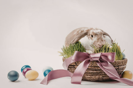 A cute Easter bunny sitting in a wicker basket with a pink bow, surrounded by colorful Easter eggs...