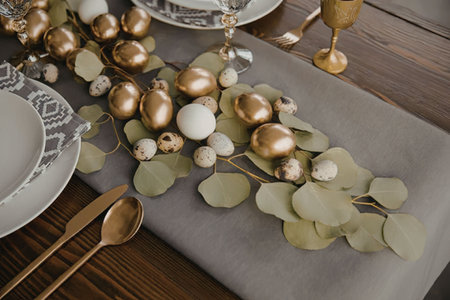 An Easter-themed table setting featuring a gray runner, gold and white eggs, green leaves, and...