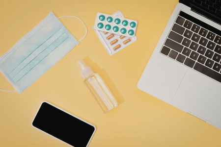 The image shows a collection of essential items for COVID-19 protection, including a face mask, hand sanitizer, pills, a smartphone, and a laptop on a yellow surface.の写真素材