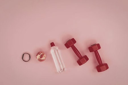 A flat lay of fitness essentials including dumbbells, a water bottle, an apple, and a hair tie on a pink background.の写真素材