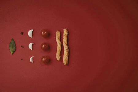 A creative arrangement of food ingredients on a red background, featuring tomatoes, mozzarella cheese, basil leaf, and breadsticks.の写真素材