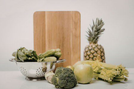 A still life of fresh produce including broccoli, apple, celery, pineapple, and leafy greens on a...