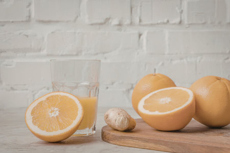 A refreshing setup of oranges, one cut open, alongside a glass of orange juice and a small ginger on a wooden board against a white brick wall.の写真素材
