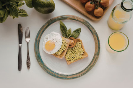 A plate of avocado toast with a fried egg, basil leaves, and cherry tomatoes on the side,...