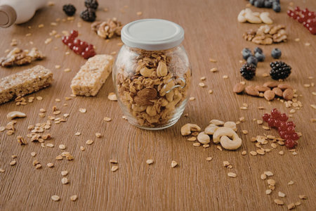 A glass jar filled with granola surrounded by various nuts and dried fruits on a wooden table.の写真素材