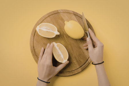 A person cutting and holding lemon halves on a wooden board with a yellow background