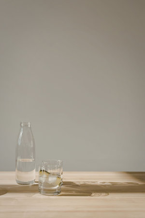 A clear glass bottle and a short glass with ice are on a wooden table against a gray background.の写真素材
