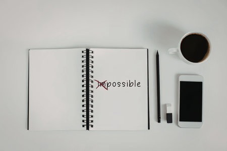 A flat lay of a notebook with the word 'impossible' and a red arrow turning it into 'possible', a pen, a smartphone, a charger and a cup of coffee on a white surface.の写真素材