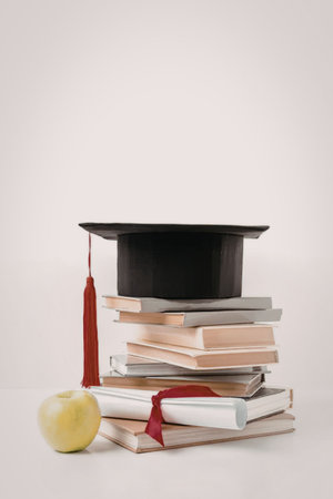 A black graduation cap sits atop a stack of books with a red tassel hanging down. A green apple and a rolled up diploma with a red ribbon are in front of the stack.の写真素材