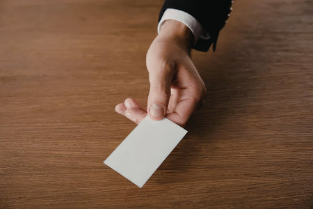 A hand in a business suit holding a blank white business card over a wooden table.の写真素材