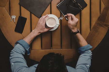 A man sitting at a wooden table, holding a cup of coffee in one hand and a pen in the other, with a...