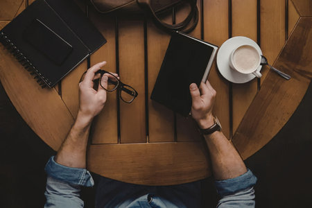 A man sitting at a wooden table, working on a tablet with a pen in hand, with a cup of coffee and a...
