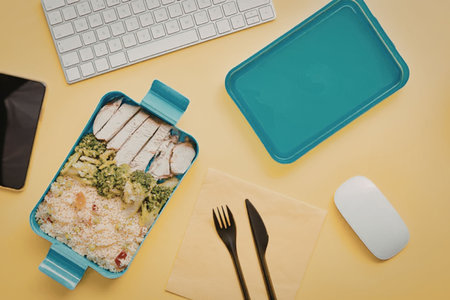 A healthy lunch setup with a blue lunch box containing salad and chicken, a keyboard, computer...