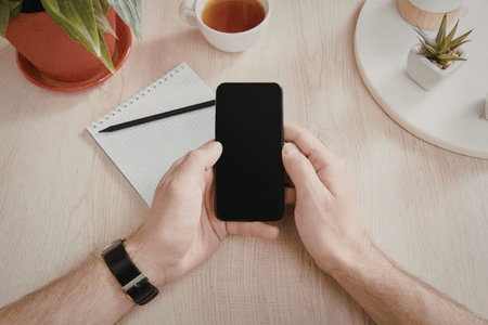 A person holding a smartphone on a desk with a notebook, pen, cup of tea, and potted plants.