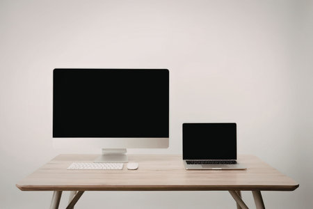 A modern workspace featuring a computer monitor and a laptop on a wooden desk against a plain white...