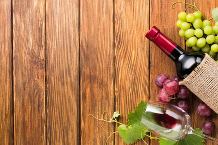 a bottle of red wine with a red foil top, a wine glass half full of red wine, and clusters of red and green grapes on a wooden table