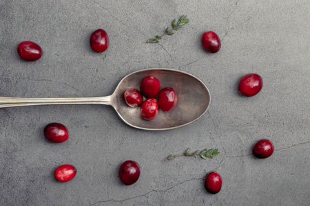 a silver spoon filled with several cranberries surrounded by more cranberries and small green leaves on a gray surface