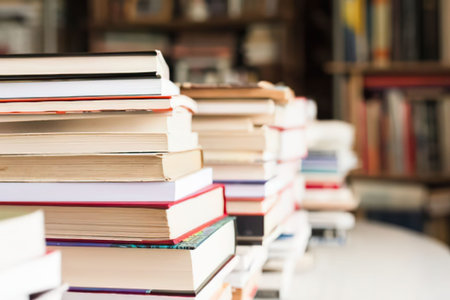 A close-up view of multiple stacks of books with colorful spines and pages facing upwards, placed on a white table in a blurred library background with shelves of books.