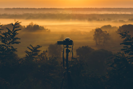 Silhouette of a camera on a tripod on a hill capturing the morning fog. Countryside landscape with a camera on a tripod, sunrise and morning fog.の写真素材
