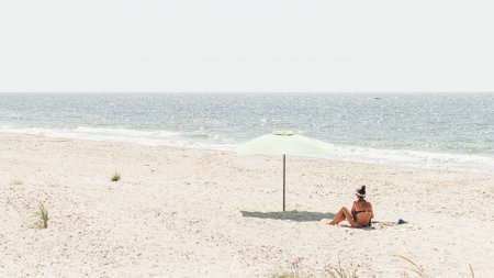 Rear view of a young woman sitting with an umbrella on the beach during a summer vacation. A young tanned woman in a swimsuit sits and relaxes on a tropical beach with white sand.の写真素材
