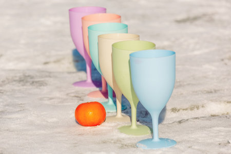 Six pastel-colored plastic wine glasses stand in a row on a snowy surface, accompanied by bright orange tangerines, creating a vivid contrast.の写真素材