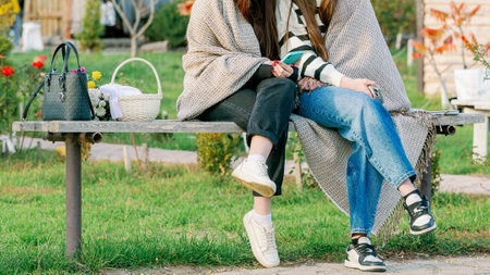 Two young women sitting together outdoors, wrapped in a cozy blanket, holding smartphones and enjoying a casual moment in cool weather.の写真素材
