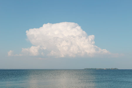 A massive cumulus cloud hovers over a tranquil blue sea with a distant island in the background, creating a peaceful scenic view.の写真素材